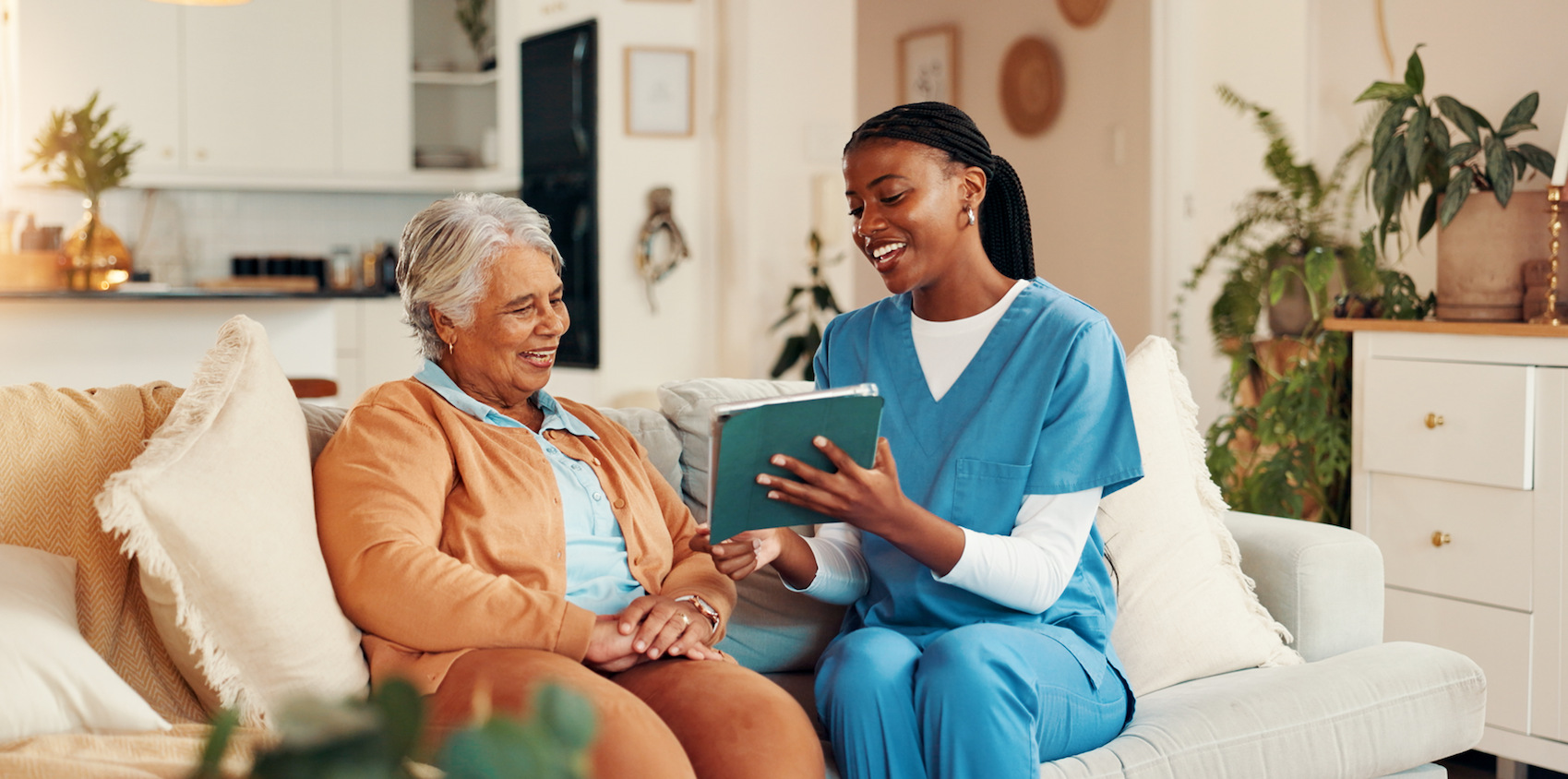 A nurse in blue scrubs sits on a couch next to an elderly woman, showing her a notebook and smiling. They appear to be having a friendly conversation in a bright, cozy living room.